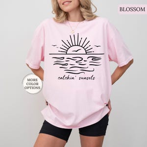 May include: Light pink t-shirt with a black graphic of a sunset over water, with the words "catchin' sunsets" below. The shirt has a relaxed fit and is made of a soft material. The words "More Color Options" are in a circle on the left.