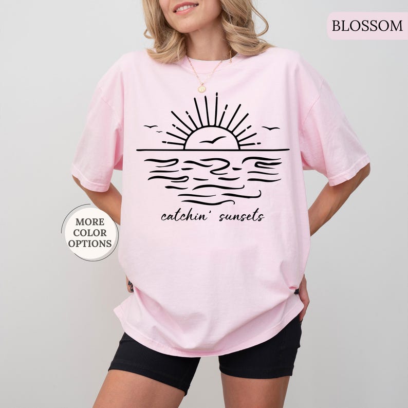May include: Light pink t-shirt with a black graphic of a sunset over water, with the words "catchin' sunsets" below. The shirt has a relaxed fit and is made of a soft material. The words "More Color Options" are in a circle on the left.
