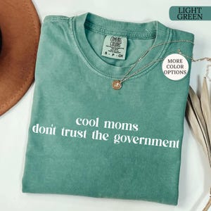 Peut inclure: T-shirt vert clair avec l'inscription blanche "cool moms don't trust the government". Le t-shirt a un col rond et des manches courtes. Un collier dor&eacute; avec un pendentif est drap&eacute; sur le t-shirt.