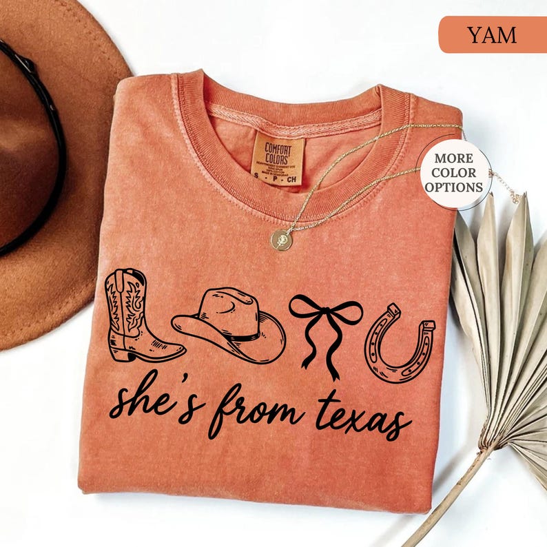 Pode incluir: Uma camiseta cor inhame com gola redonda e mangas curtas. A camisa apresenta um gr&aacute;fico preto de uma bota de cowboy, chap&eacute;u, la&ccedil;o e ferradura, com o texto "she's from texas" em uma fonte cursiva.