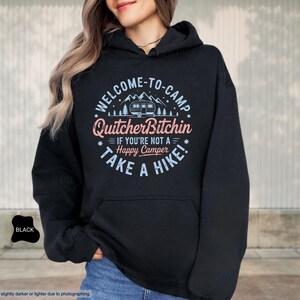 May include: Black hooded sweatshirt with the text "WELCOME-TO-CAMP Quitcher Bitchin IF YOU'RE NOT A Happy Camper TAKE A HIKE!" in a circular design. The design includes a camper, trees, and stars. The sweatshirt has a front pocket and a drawstring hood.