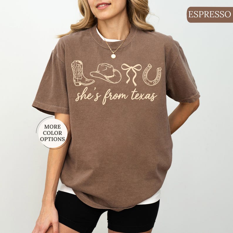 Pode incluir: Camiseta cor de caf&eacute; com um gr&aacute;fico branco com uma bota de cowboy, chap&eacute;u, la&ccedil;o e ferradura, com o texto "she's from texas". A camiseta tem mangas curtas e decote redondo.