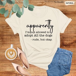May include: Cream-colored t-shirt with the text "apparently I'm not allowed to adopt All the dogs - rude, but okay." The shirt is paired with blue jeans and blush-colored heels. A cup of coffee with latte art is also visible.