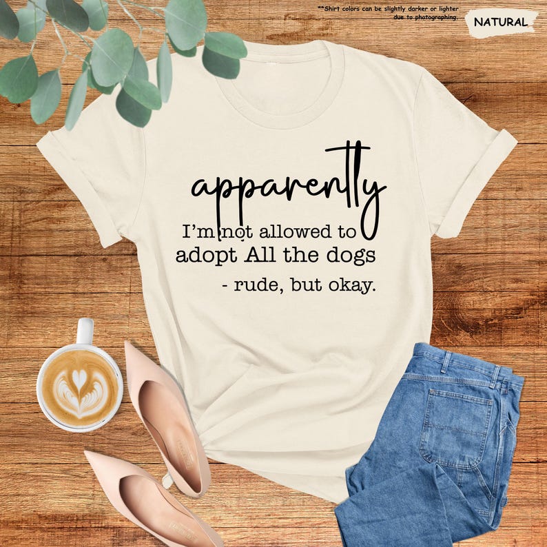 May include: Cream-colored t-shirt with the text "apparently I'm not allowed to adopt All the dogs - rude, but okay." The shirt is paired with blue jeans and blush-colored heels. A cup of coffee with latte art is also visible.
