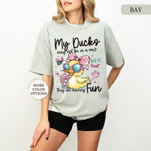 May include: A light green t-shirt with the text "My Ducks may not be in a row but at least they are having Fun." The shirt features a cartoon duck wearing sunglasses and a pink bow, holding a drink.