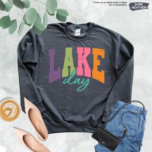 Op de afbeelding: Donkergrijs sweatshirt met de tekst "LAKE day" in kleurrijke blokletters. De afbeelding bevat een spijkerbroek, een zwarte handtas en nude-kleurige schoenen. Een kopje koffie met latte art is ook zichtbaar.
