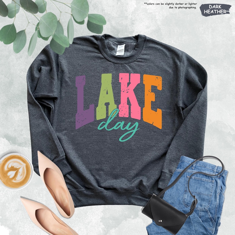 Op de afbeelding: Donkergrijs sweatshirt met de tekst "LAKE day" in kleurrijke blokletters. De afbeelding bevat een spijkerbroek, een zwarte handtas en nude-kleurige schoenen. Een kopje koffie met latte art is ook zichtbaar.