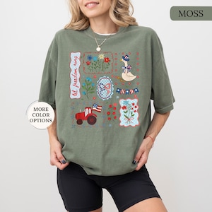 May include: Moss green t-shirt with patriotic-themed embroidered patches. Patches include flowers, a goose in a hat, a tractor with a flag, and the text "let freedom ring". The shirt has a relaxed fit.
