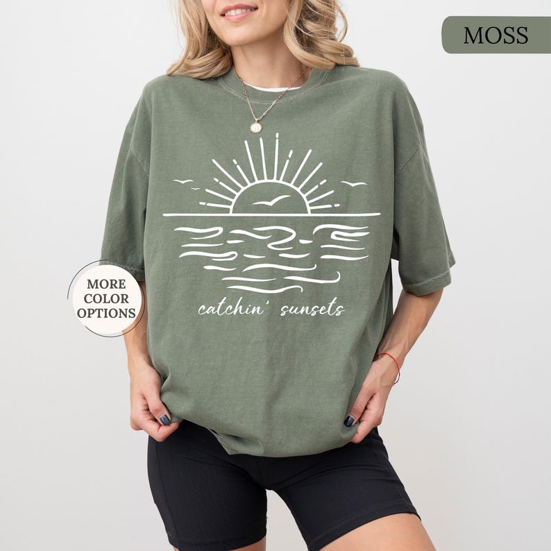 May include: Moss green t-shirt with a white sunset graphic. The design includes a sun, waves, and the text "catchin' sunsets". The shirt has short sleeves and a relaxed fit. Additional color options are available.