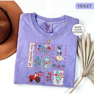 May include: A violet Comfort Colors t-shirt with a patriotic design. The design includes a red tractor, flowers, a goose wearing a hat, and the text "let freedom ring". A gold necklace is draped over the shirt.