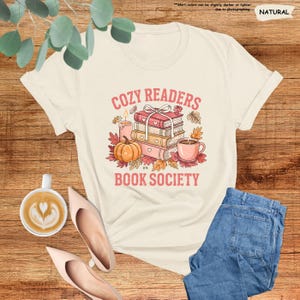 May include: Cream-colored t-shirt with a graphic that reads "COZY READERS BOOK SOCIETY." The design features a stack of books, a pumpkin, a cup of coffee, and autumn leaves. The shirt is paired with blue jeans and pink shoes.