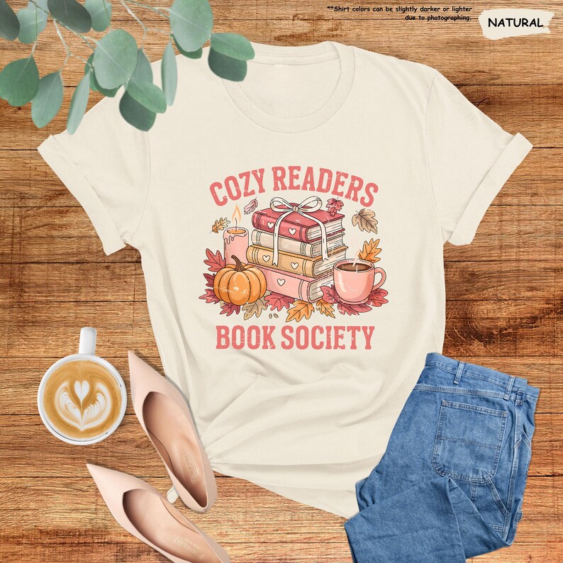 May include: Cream-colored t-shirt with a graphic that reads "COZY READERS BOOK SOCIETY." The design features a stack of books, a pumpkin, a cup of coffee, and autumn leaves. The shirt is paired with blue jeans and pink shoes.