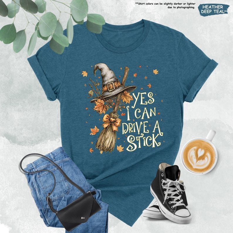 Pode incluir: Camiseta azul-petr&oacute;leo com um desenho de chap&eacute;u de bruxa e vassoura, o texto diz "YES I CAN DRIVE A STICK". A camiseta &eacute; combinada com jeans azuis, uma bolsa preta e t&ecirc;nis de cano alto pretos. Uma x&iacute;cara de caf&eacute; tamb&eacute;m est&aacute; na imagem.