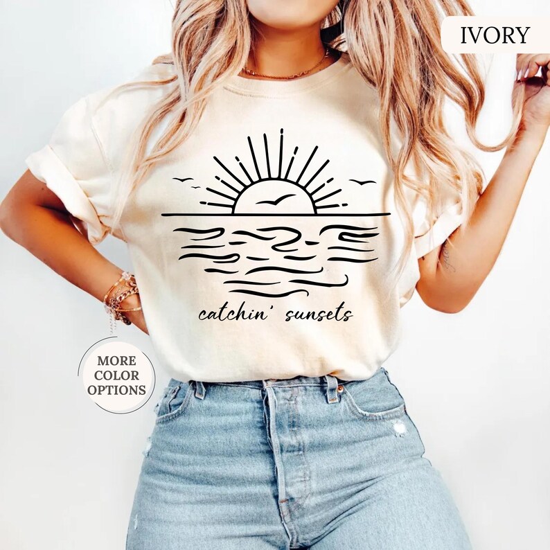 May include: Ivory-colored t-shirt featuring a black graphic of a sunset over the ocean with the text "catchin' sunsets". The design includes a sun, birds, and waves. The shirt has rolled-up sleeves.