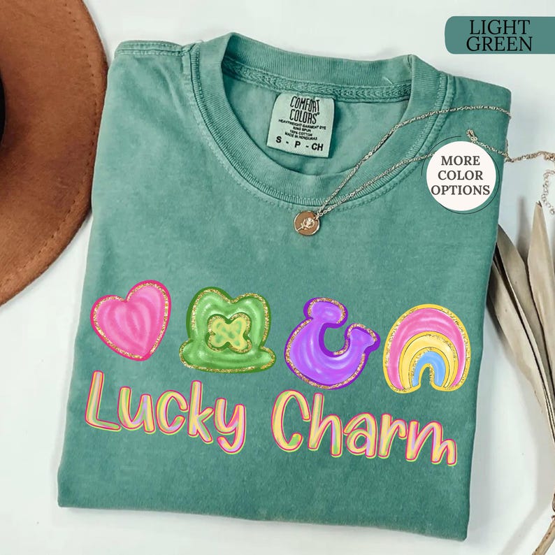 May include: Light green Comfort Colors t-shirt with a colorful "Lucky Charm" graphic. The graphic includes a pink heart, green shamrock, purple ampersand, and a rainbow. A silver necklace with a pendant is on the shirt.