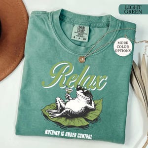 May include: Light green t-shirt with a graphic of a frog wearing sunglasses, holding a cocktail, and lounging on a lily pad. The word "Relax" is above the frog, and "Nothing is Under Control" is below.