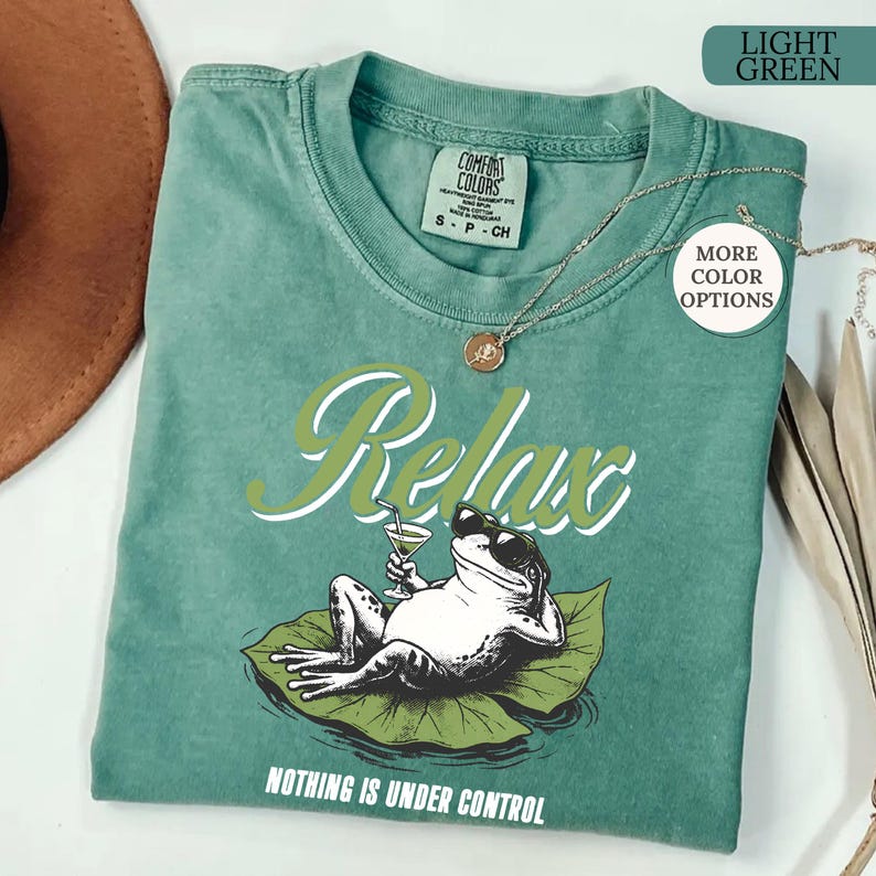May include: Light green t-shirt with a graphic of a frog wearing sunglasses, holding a cocktail, and lounging on a lily pad. The word "Relax" is above the frog, and "Nothing is Under Control" is below.
