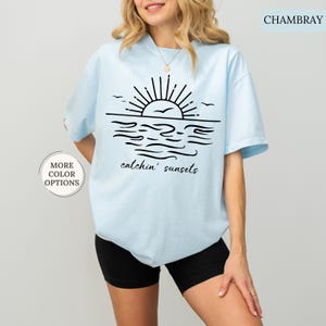 May include: Light blue t-shirt with a black graphic of a sunset over the ocean, including the text "catchin' sunsets". The shirt has short sleeves and a round neckline. The words "More Color Options" are in a circle on the left side of the image.
