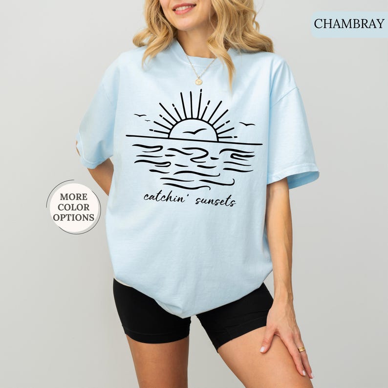 May include: Light blue t-shirt with a black graphic of a sunset over the ocean, including the text "catchin' sunsets". The shirt has short sleeves and a round neckline. The words "More Color Options" are in a circle on the left side of the image.