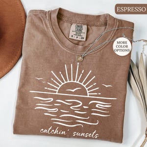 May include: Espresso-colored t-shirt with a white graphic of a sunset over the ocean, with the words "catchin' sunsets" below. The shirt has a crew neck and short sleeves. A silver necklace with a pendant is draped over the shirt.