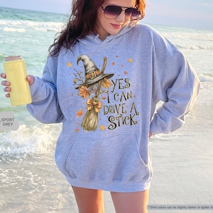 Op de afbeelding: Een lichtgrijze hoodie met een heksenthema. De afbeelding bevat een heksenhoed, bezem en de tekst "YES I CAN DRIVE A STICK". De hoodie is sportgrijs van kleur.