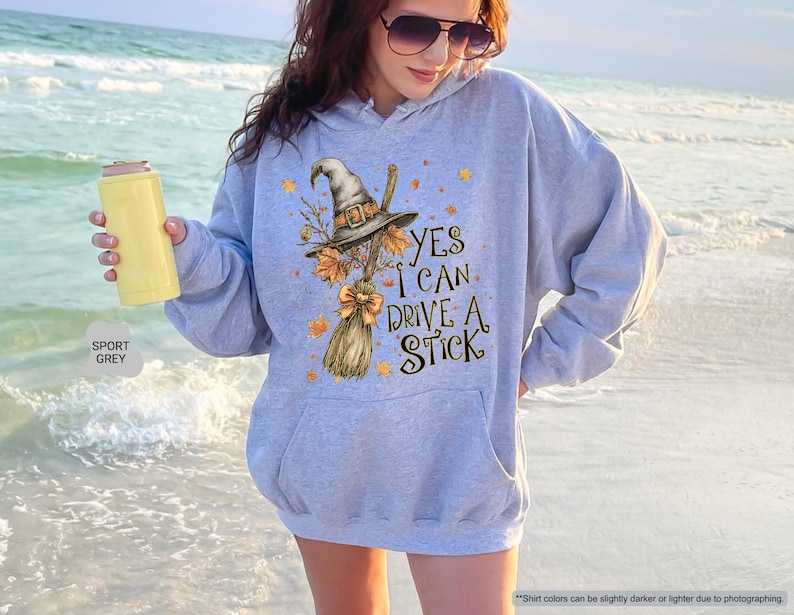 Op de afbeelding: Een lichtgrijze hoodie met een heksenthema. De afbeelding bevat een heksenhoed, bezem en de tekst "YES I CAN DRIVE A STICK". De hoodie is sportgrijs van kleur.