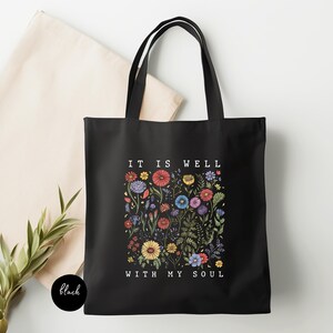 May include: Black tote bag with the text "IT IS WELL WITH MY SOUL" and a floral design in various colors. The bag has two black handles and is made of a sturdy fabric. A beige tote bag is visible in the background.