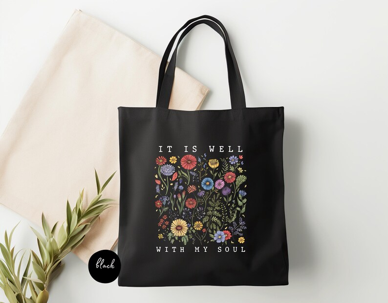 May include: Black tote bag with the text "IT IS WELL WITH MY SOUL" and a floral design in various colors. The bag has two black handles and is made of a sturdy fabric. A beige tote bag is visible in the background.