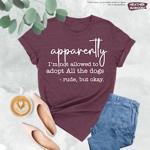 May include: Heather maroon t-shirt with the text "apparently I'm not allowed to adopt All the dogs - rude, but okay." The shirt is paired with blue jeans, beige flats, and a cup of coffee.