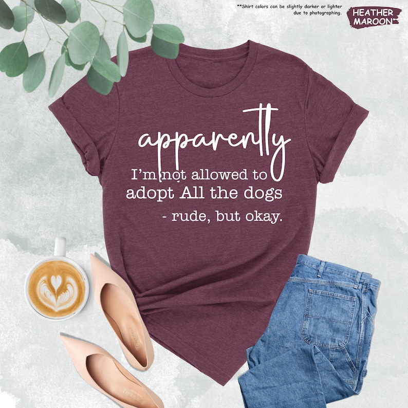 May include: Heather maroon t-shirt with the text "apparently I'm not allowed to adopt All the dogs - rude, but okay." The shirt is paired with blue jeans, beige flats, and a cup of coffee.