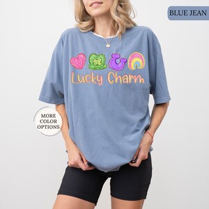 May include: Blue jean colored t-shirt with a graphic of a pink heart, green shamrock, purple horseshoe, and rainbow. The text "Lucky Charm" is written in yellow. The shirt is a casual style.