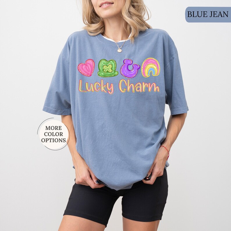 May include: Blue jean colored t-shirt with a graphic of a pink heart, green shamrock, purple horseshoe, and rainbow. The text "Lucky Charm" is written in yellow. The shirt is a casual style.