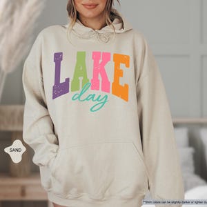 Op de afbeelding: Zandkleurige hoodie met een voorzak en een capuchon met trekkoord. De voorkant heeft de tekst "LAKE day" in paars, groen, roze, oranje en turquoise. De hoodie heeft een relaxte pasvorm.