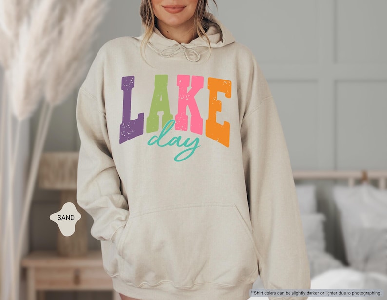 Op de afbeelding: Zandkleurige hoodie met een voorzak en een capuchon met trekkoord. De voorkant heeft de tekst "LAKE day" in paars, groen, roze, oranje en turquoise. De hoodie heeft een relaxte pasvorm.