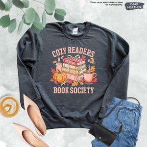 May include: Dark heather gray sweatshirt with the text "COZY READERS BOOK SOCIETY" in a pink and orange design. The graphic includes a stack of books, a pumpkin, a cup of coffee, and autumn leaves.