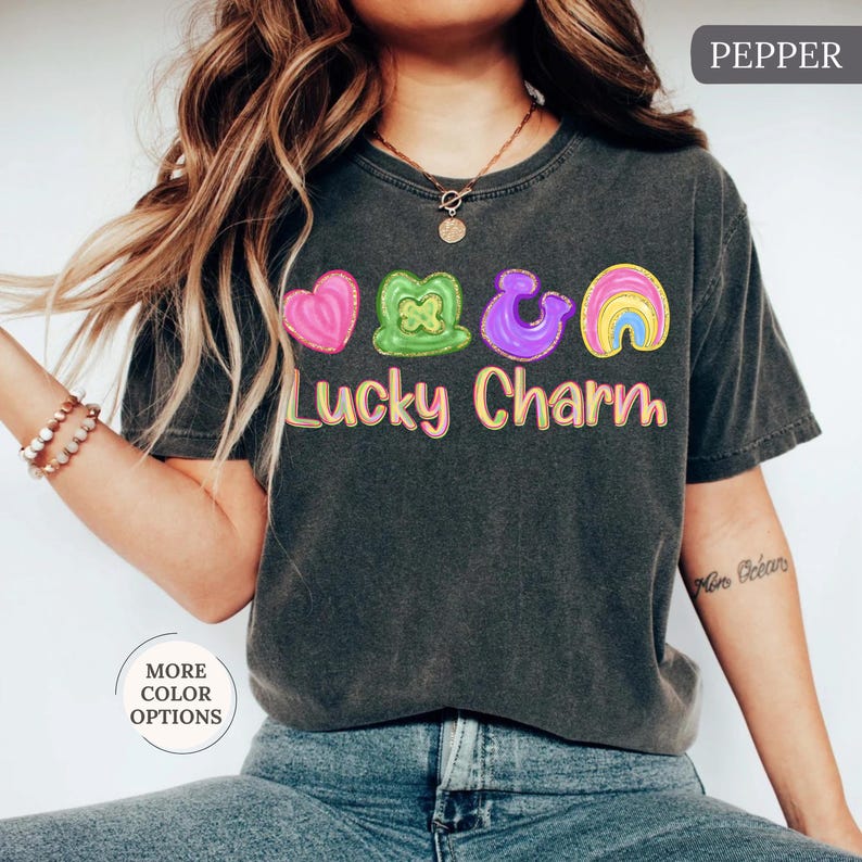 May include: A dark gray t-shirt with a colorful "Lucky Charm" graphic. The graphic features a pink heart, green shamrock, purple horseshoe, and a rainbow. The shirt has short sleeves and a crew neck.