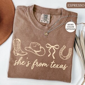 Pode incluir: Camiseta cor de caf&eacute; com tema western. A camiseta apresenta um gr&aacute;fico de bota, chap&eacute;u de cowboy, la&ccedil;o e ferradura, com o texto "she's from texas" em fonte cursiva. Um colar de prata est&aacute; drapeado sobre a camiseta.