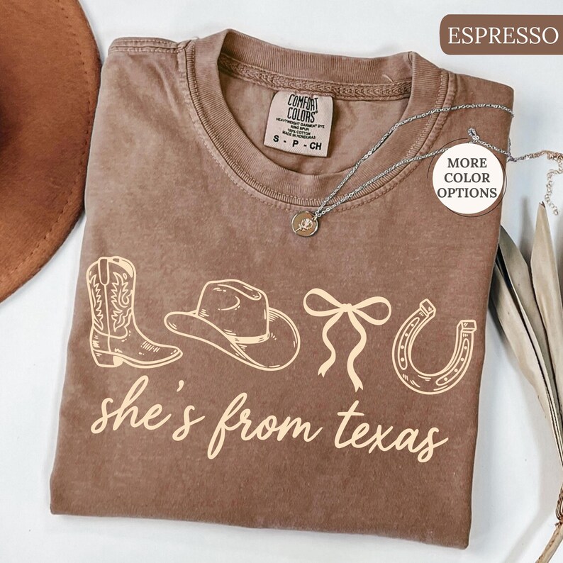 Pode incluir: Camiseta cor de caf&eacute; com tema western. A camiseta apresenta um gr&aacute;fico de bota, chap&eacute;u de cowboy, la&ccedil;o e ferradura, com o texto "she's from texas" em fonte cursiva. Um colar de prata est&aacute; drapeado sobre a camiseta.