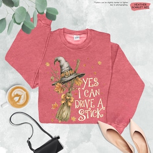 Op de afbeelding: Heather scharlakenrood sweatshirt met een heksenhoed, bezem en herfstbladeren grafisch. De tekst luidt "Yes I Can Drive A Stick". Een leuke top voor Halloween of de herfst.