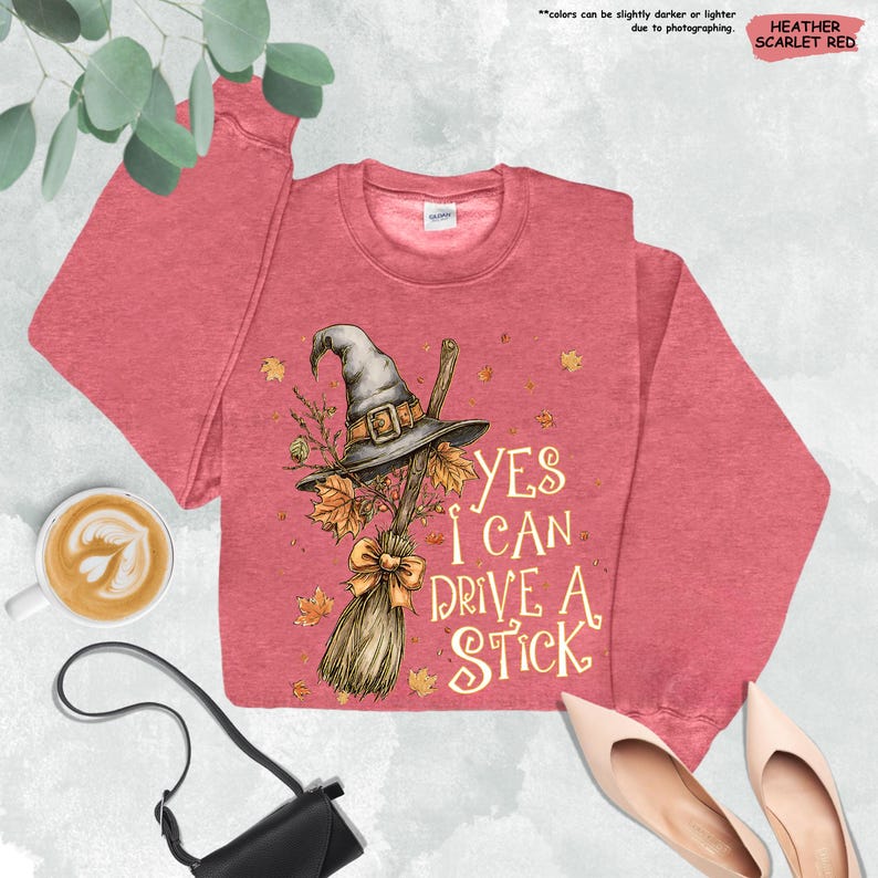 Op de afbeelding: Heather scharlakenrood sweatshirt met een heksenhoed, bezem en herfstbladeren grafisch. De tekst luidt "Yes I Can Drive A Stick". Een leuke top voor Halloween of de herfst.