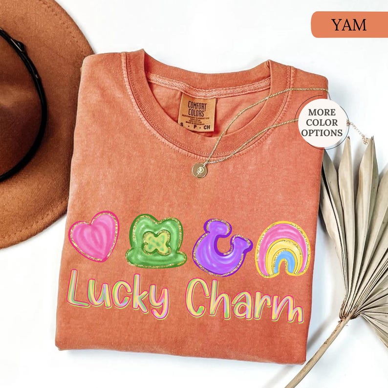 May include: A yam-colored t-shirt with a "Lucky Charm" graphic. The graphic includes a pink heart, a green shamrock, a purple ampersand, and a rainbow. A gold necklace is draped over the shirt.