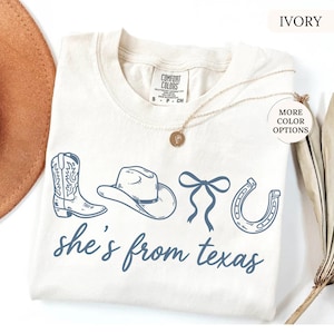 Pode incluir: Camiseta cor marfim com um gr&aacute;fico azul de uma bota de cowboy, chap&eacute;u, la&ccedil;o e ferradura. O texto "she's from texas" est&aacute; escrito em azul. Um colar de ouro est&aacute; sobre a camiseta.