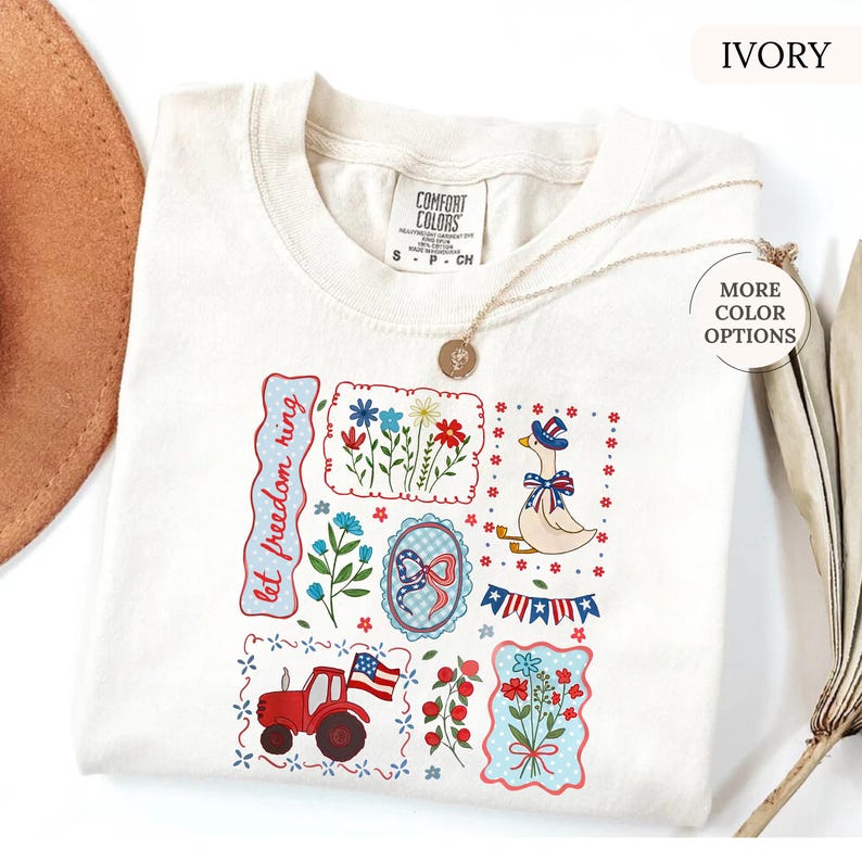 May include: Ivory-colored Comfort Colors t-shirt with a patriotic design. The design includes a red tractor, flowers, a goose in a hat, and the text "let freedom ring". A gold necklace is draped over the shirt.