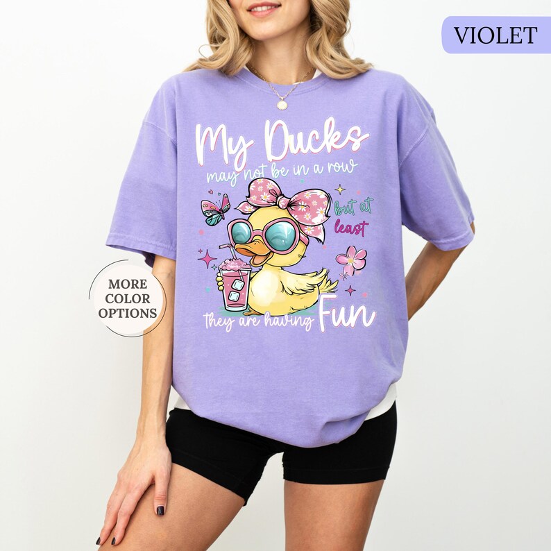 May include: A violet t-shirt with a graphic of a yellow duck wearing sunglasses and a pink bow, holding a drink. The text reads "My Ducks may not be in a row but at least they are having Fun". "More Color Options" is also displayed.