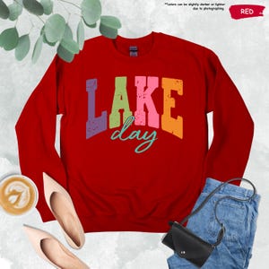 Op de afbeelding: Een rode sweatshirt met de tekst "LAKE day" in kleurrijke, verweerde letters. De sweatshirt wordt getoond met een paar beige schoenen, een kopje koffie, een zwarte handtas en een spijkerbroek.