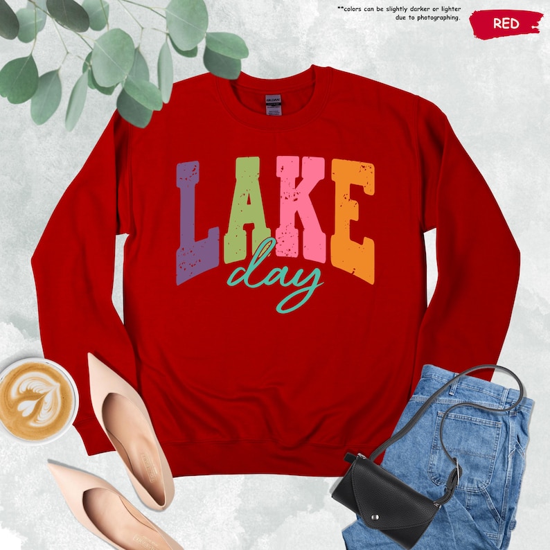 Op de afbeelding: Een rode sweatshirt met de tekst "LAKE day" in kleurrijke, verweerde letters. De sweatshirt wordt getoond met een paar beige schoenen, een kopje koffie, een zwarte handtas en een spijkerbroek.