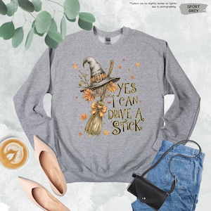 Pode incluir: Camisola cinzenta desportiva com um chap&eacute;u de bruxa, uma vassoura e folhas de outono. O texto "YES I CAN DRIVE A STICK" est&aacute; impresso na frente. Gola redonda.