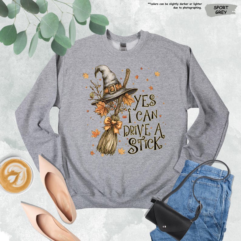 Pode incluir: Camisola cinzenta desportiva com um chap&eacute;u de bruxa, uma vassoura e folhas de outono. O texto "YES I CAN DRIVE A STICK" est&aacute; impresso na frente. Gola redonda.