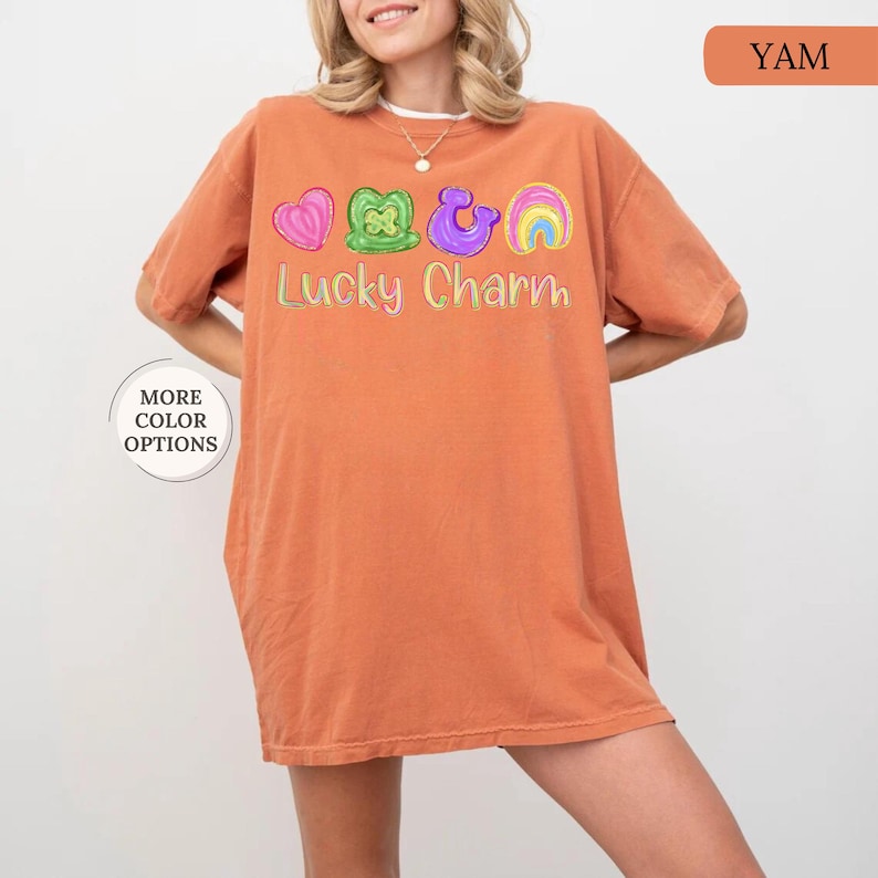 May include: An orange-colored t-shirt with the words "Lucky Charm" embroidered in a colorful design. The design includes a pink heart, green shamrock, purple horseshoe, and a rainbow. The shirt has short sleeves and a relaxed fit.