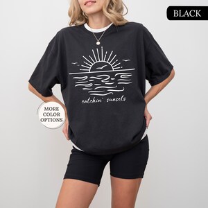 May include: Black short-sleeved t-shirt with a white graphic of a sunset over the ocean, with the words "catchin' sunsets" below. The shirt has a relaxed fit and is worn with black shorts. The words "MORE COLOR OPTIONS" are in a white circle.